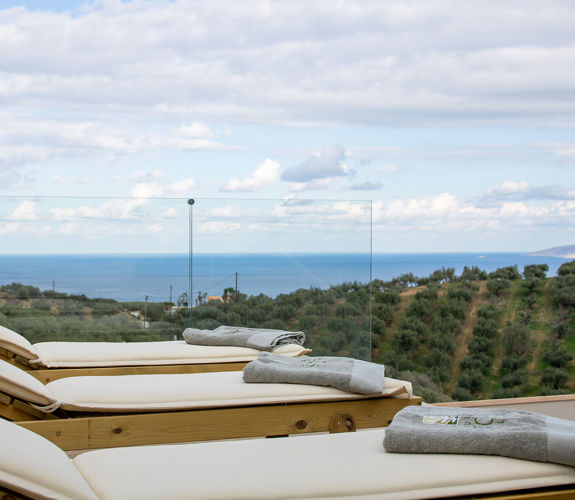 Sunbeds with towels overlooking olive groves and the sea, behind a glass terrace railing