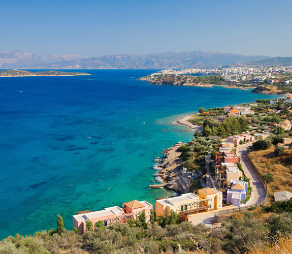 Panoramic coastal view with colorful buildings, blue waters, and distant mountains in Crete